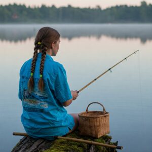 A girl in a blue camp shirt fishing quietly by the lakeside with a basket beside her.