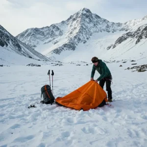 A person setting up a compact avalanche backpack tent in a snowy mountain environment.