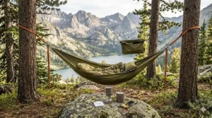 A hiker relaxing in a portable camping hammock suspended above a peaceful mountain lake during a backpacking trip.