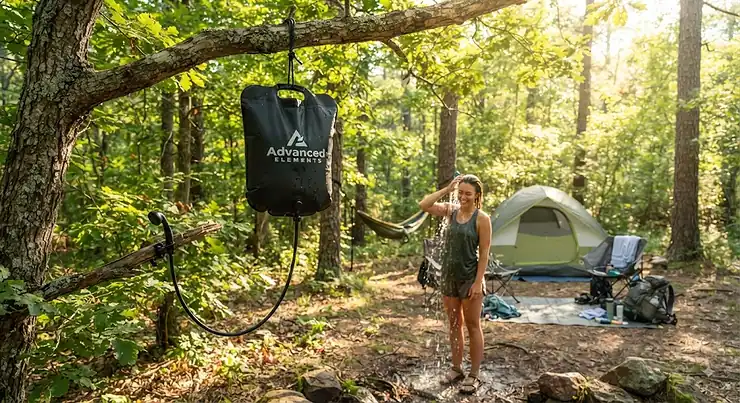 A portable camp shower bag hanging from a tree branch at a campsite during a sunny afternoon.